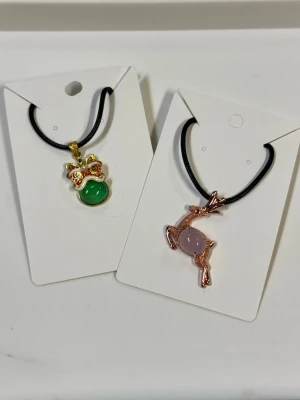 Två halsband med jade djurmotiv och pärlor - Säljer två halsband med svarta band och unika hängen. Ett halsband har ett guld- och rött dekorerat hänge med grön jade, det andra har ett roséguldfärgat hjorthänge med en ljuslila jade. Perfekta för dig som gillar söta accessoarer med djurtema.