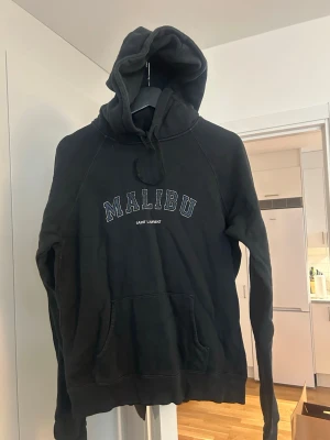 Svart hoodie Saint Laurent Malibu - Säljer en svart hoodie från Saint Laurent med 'MALIBU' tryck på bröstet. Storlek M men liten i storlek så passar S snarare. Använd fåtal ggr så fint skick förutom märkeslappen som sitter löst (se bild 4). Pris kan diskuteras 