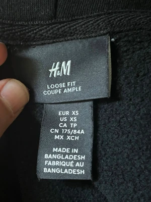 Svart hoodie med dragkedja från H&M - Säljer en svart hoodie från H&M i loose fit-modell. Hoodien har dragkedja framtill, huva med dragsko och två fickor på sidorna. Perfekt för en avslappnad och stilren look. Tillverkad i mjukt material som känns skönt mot huden.