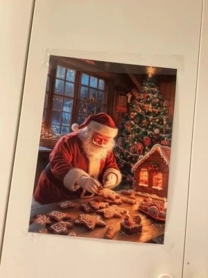 Julposter med tomte och pepparkakor - Säljer en färgglad poster med julmotiv där en tomte dekorerar pepparkakor vid ett bord. I bakgrunden syns en pyntad julgran och ett pepparkakshus. Postern har varma färger som rött, grönt och brunt och är tryckt på papper. 30X40cm 