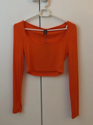 Orange kort croppad topp - Snygg orange kort croppad topp med lång ärm och rundad urringning. Tillverkad i ett stretchigt syntetmaterial som sitter snyggt och bekvämt. Helt ny. Jättefin med blåa jeans till! Gåva vid köp🎁