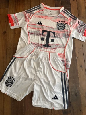 FC Bayern München matchställ  - Säljer ett FC Bayern München matchställ från Adidas i vitt med röda och grå detaljer. Tröjan har klubbmärke, sponsorlogga och Adidas-logga, samt korta ärmar. Shortsen har elastisk midja, klubbmärke och klassiska Adidas-ränder. Perfekt för fotbollsträning eller match.