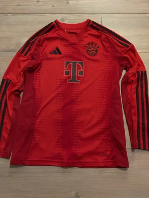 Röd Bayern München tröja Adidas - Snygg röd långärmad matchtröja från FC Bayern München med svarta detaljer och tryck. Adidas-logga på bröstet, klubbmärke och sponsortryck fram. Nummer 6 och Kimmich på ryggen. Tillverkad i funktionsmaterial, perfekt för fotbollsträning.