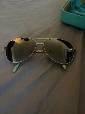 Silvriga aviator solglasögon - Snygga aviator solglasögon med silvrig metallbåge och mörka tonade glas. Klassisk pilotform med näskuddar och tunna skalmar. Perfekta för dig som vill ha en tidlös och cool look. Kommer med ett turkost fodral.