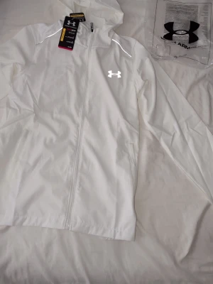 Vit vindjacka från Under Armour - Snygg vit vindjacka från Under Armour med huva och hel dragkedja framtill. Jackan har reflekterande logga på bröstet och stor reflekterande text på ryggen. Tillverkad i lätt och tåligt material, perfekt för blåsiga dagar. Två sidofickor med dragkedja.