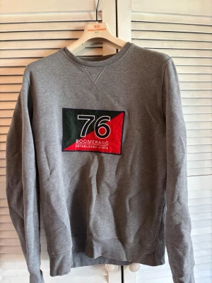 Grå sweatshirt från Boomerang - Grå sweatshirt med stort broderat tryck på bröstet i svart, vitt, rött och grönt med texten '76 Boomerang Established in 1976'. Klassisk rund halsringning och långa ärmar. Perfekt för en avslappnad och stilren look.