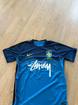 Blå Brasilien fotbollströja Nike x Stüssy - Snygg blå fotbollströja från Nike i samarbete med Stüssy och Brasilien. Tröjan har korta ärmar, gul Nike-logga, Brasilien-emblem och Stüssy-tryck på bröstet. Tillverkad i Dri-FIT material som andas, perfekt för träning eller streetwear. Hör gärna av er vid frågor och pris kan diskuteras 