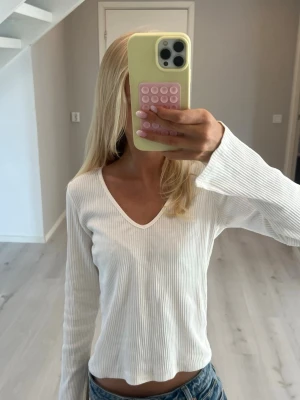 Vit ribbad långärmad topp - Säljer en enkel och stilren vit ribbad topp med v-ringning och långa ärmar. Toppen har en lätt croppad passform och är gjord i ett mjukt, stretchigt material som sitter snyggt på kroppen. Armarna är slitsade och öppna. Perfekt att matcha med jeans eller kjol. ❤️❤️❤️ (lappar är bortklippta)