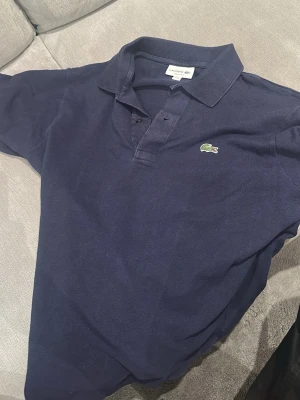 Marinblå pikétröja från Lacoste - Klassisk marinblå pikétröja från Lacoste med den ikoniska krokodilloggan på bröstet. Tröjan har krage, knappar framtill och är tillverkad i bomull. Perfekt för en stilren och avslappnad look.