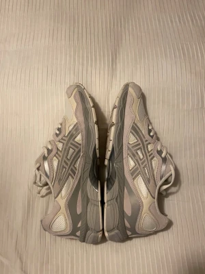 Asics Gel-NYC Cream Cream - Snygga sneakers från Asics i vitt och ljusgrått med klassiska mesh- och syntetpaneler. Skorna har den ikoniska Asics-loggan på sidan och en chunky sula som ger retrovibbar. Perfekta för dig som gillar sportig och stilren look.