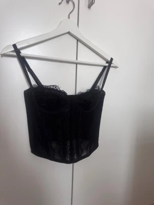 Svart spetskorsett med tunna axelband - Säljer en svart korsettopp med tunna axelband och genombruten spets. Toppen har croppad passform och är tillverkad i polyester och syntetmaterial. Perfekt för att skapa en trendig och edgy look.