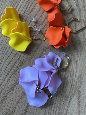 Färgglada blommiga örhängen - Unika örhängen i form av stora blommor i plast, finns i lila, gul och orange. Hängena har vågiga blomblad och sitter på guldfärgade ringar. Perfekta för dig som vill sticka ut med färg och cool design.
