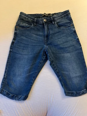 Blå jeansshorts från Name It - Snygga blå jeansshorts från Name It med klassisk femficksdesign och knappgylf. Shortsens denim har en lätt tvättad look och raka ben som slutar strax ovanför knäna. Perfekta för en avslappnad stil under varmare dagar.