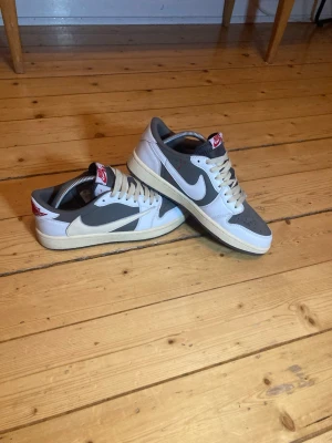 Nike Air Jordan x travis 1 Low  - Säljer ett par Nike Air Jordan 1 Low i brunt och vitt med klassisk swoosh på sidan. Skorna har beige sula, grått tåparti och vita paneler. Röd logga på plösen och hälen. Tillverkade i läder och textil, perfekta för en clean streetwear-look.