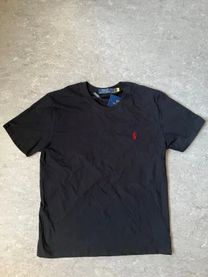Polo Ralph Lauren svart t-shirt - Klassisk svart t-shirt från Polo Ralph Lauren med korta ärmar och rund hals. T-shirten har den ikoniska röda loggan broderad på bröstet och är gjord i mjuk bomull för en skön känsla. Perfekt basplagg till garderoben.