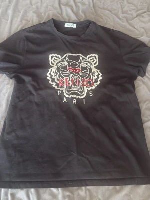 Svart Kenzo t-shirt med tigertryck - Svart t-shirt från Kenzo med ikoniskt broderat tigerhuvud och texten 'KENZO PARIS' i vitt och rött på bröstet. Klassisk passform och rund hals. Perfekt statement-plagg för dig som gillar streetwear och snygga detaljer.
