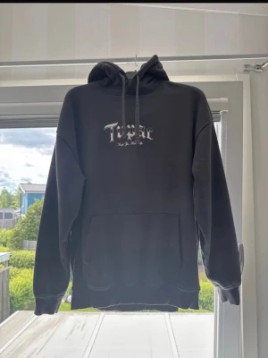 Svart Tupac hoodie med tryck - Säljer en svart hoodie med stor Tupac-tryck på både fram- och baksidan. Hoodien har huva med dragsko och en stor magficka. Texten 'Tupac' och 'Keep Ya Head Up' syns tydligt i vitt. Perfekt för dig som gillar streetwear och hiphopstil.