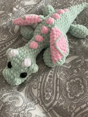 Virkad drake  - Supergullig handvirkad drake i mintgrönt garn med rosa detaljer på ryggen och vingarna samt vita horn. Draken har stora svarta ögon och är gjord i mjukt, fluffigt material. Perfekt som prydnad eller gosedjur.