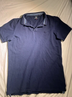 Marinblå Superdry Piké strl M - Klassisk mörkblå pikétröja från Superdry med korta ärmar och diskret broderad logga på bröstet. Tröjan har krage med knappslå och vita detaljer på insidan av kragen. Perfekt till jeans eller shorts för en clean look. Fraktar snabbt och öppen för rimliga bud.
