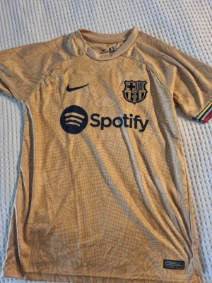 Beige FC Barcelona matchtröja Nike - Säljer en beige FC Barcelona matchtröja från Nike med mörkblå logga och Spotify-tryck på bröstet. Tröjan har korta ärmar, rund hals och ett diskret kartmönster över hela tyget. Perfekt för fotbollsträning eller att bära som supporter.