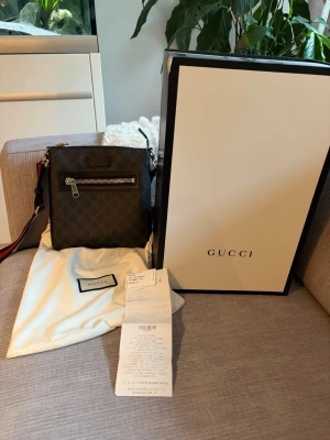 Gucci axelväska med monogram - Snygg axelväska från Gucci i mörkgrått med klassiskt GG-monogram. Väskan har en silverfärgad dragkedja framtill, justerbar axelrem med röd och blå rand samt innerfack för småprylar. Tillbehör som dustbag och originalkartong medföljer.