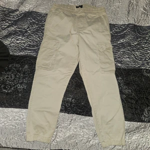 Beige cargopants från Pull&Bear - Säljer ett par beige cargopants från Pull&Bear med elastisk midja och snörning. Byxorna har flera praktiska fickor på sidorna och baktill, samt resår vid bensluten för en chill och avslappnad look. Perfekt för dig som gillar streetwear och funktionella detaljer.