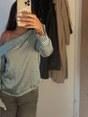 Randig offshoulder blus - Snygg offshoulder blus med svarta och vita vertikala ränder. Blusen har långa ärmar med breda manschetter och en bred krage som går över ena axeln. Tillverkad i ett lätt lite silkigt material som ger en stilren och trendig look.