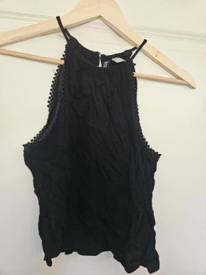 Svart ärmlös linnetopp H&M 32 - Säljer en svart ärmlös linnetopp från H&M med smala axelband och dekorativa små bollar längs kanterna. Toppen har en lätt rynkad struktur och hög halsringning. Perfekt att styla med jeans eller kjol.