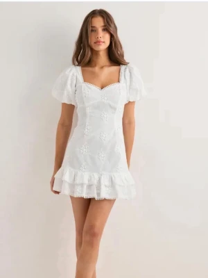 Vit kort klänning med rosett - Supersöt vit kort klänning med broderade blommor, puffärmar och volangkant. Klänningen har öppen rygg med stor rosett och fina spetsdetaljer längs kanterna. Perfekt för sommarens alla tillfällen! Prislappen är fortfarande kvar och är endast testad. 