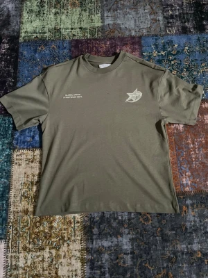 Olivgrön t-shirt från primark  - Olivgrön t-shirt med vit tryckt logga och text på både bröstet och ryggen. Oversized passform med korta ärmar och rund hals. Perfekt för dig som gillar streetwear och vill sticka ut med en unik design.