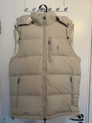 Ralph Lauren Väst Beige - Snygg beige dunväst från Ralph Lauren och har en justerbar luva. Vill sälja den ASAP så priset går att diskutera vid snabbaffär. Fråga om mer bilder om det behövs!
