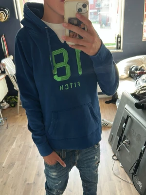 Blå hoodie från Fitch med tryck - Snygg blå hoodie från Fitch med stort grönt tryck av siffran 18 och texten 'FITCH' på bröstet. Hoodien har huva med dragsko och en stor magficka framtill. Perfekt för en avslappnad och cool stil. Materialet känns mjukt och bekvämt.