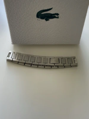 Silverfärgad klocklänk från Lacoste - Snygg silverfärgad klocklänk från Lacoste med klassisk länkdesign och diskreta mönstrade detaljer på länkarna. Tillverkad i metall och passar perfekt till din Lacoste-klocka om du vill byta ut armbandet.