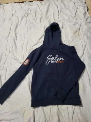 Mörkblå Sälen Ski Resort hoodie - Mörkblå hoodie med vit och orange Sälen Ski Resort-tryck på bröstet och broderad patch på ärmen med texten 'Mountain Lifestyle'. Klassisk känguruficka och huva. Perfekt för dig som gillar skidåkning och fjällkänsla.