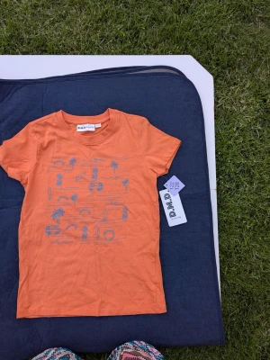 NY tshirt strl 122/128 - Orange t-shirt från D.M.D med blått tryck av palmer, bilar och surfbrädor. Klassisk rund hals och korta ärmar. Tillverkad i mjuk bomull, perfekt för en avslappnad stil.