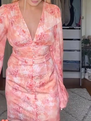 Adoore kläningen Mini - Söt klänning i rosa och koralltoner med blommigt mönster och v-ringning. Figurnära passform med dekorativa knappar framtill och långa ärmar. Perfekt för dig som gillar romantisk och trendig stil.