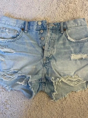 Ljusblå jeansshorts ONLY - Säljer ett par ljusblå jeansshorts från ONLY med slitna detaljer och fransig kant. Shortsen har knappar framtill, klassiska fem fickor och är tillverkade i bomull. Perfekta för sommaren och ger en avslappnad vibe.