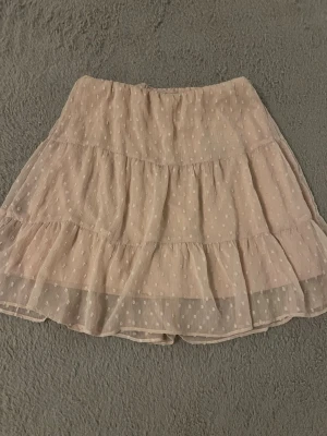 Beige volangkjol med prickigt mönster - Söt beige kortkjol med volanger och prickigt mönster. Kjolen är gjord i ett lätt och transparent material med flera lager och har en luftig känsla. Perfekt för dig som gillar en romantisk och trendig stil. Det är en avklippt klänning.