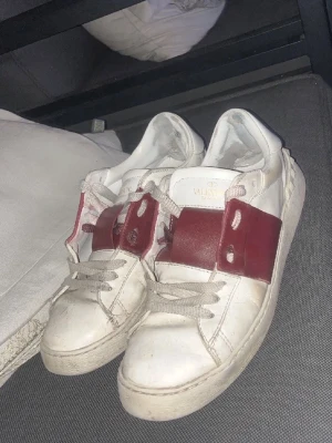 Valentino vita sneakers med röd detalj 40 - Snygga vita sneakers från Valentino med bred röd läderdetalj över snörningen. Klassisk låg modell med vita skosnören och diskret logga på plösen. Perfekta för dig som vill ha en stilren men ändå unik look. Har lite små märken som i hälen men inget som syns under användningen och små defekter 