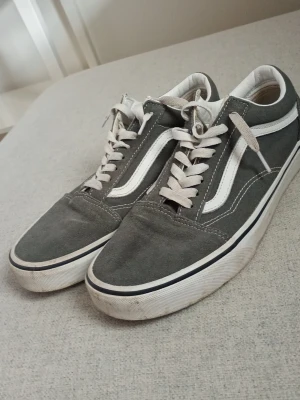 Gråa Vans Old Skool sneakers - Klassiska Vans Old Skool sneakers i grått med vit sula och vit sidostripes. Skorna har lågt skaft, vita snören och detaljer i mocka och canvas. Perfekta för en avslappnad streetwear-look.