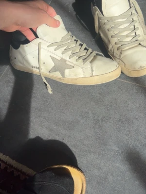 Golden Goose  - Säljer ett par vita Golden Goose sneakers med den ikoniska grå stjärnan på sidan och svarta detaljer vid hälen. Skorna har beige sula och grå skosnören. Klassisk low-top modell i läder som passar perfekt till streetwear och casual outfits.