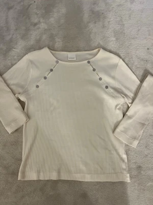 Vit ribbad topp med knappar - Stilren vit ribbad topp från Zara  med dekorativa knappar längs axlarna. Toppen har trekvartsärm och en mjuk, stretchig känsla som passar perfekt till många olika outfits. Enkel och snygg design som är lätt att matcha.