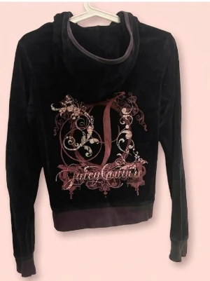 Svart velour hoodie Juicy Couture - Svart velour hoodie från Juicy Couture med dragkedja framtill och kängurufickor. Stor dekorativ logga i rosa och silver på ryggen. Huva med kantband och ribbade muddar vid ärmar och nederkant. Perfekt för en avslappnad och trendig look.