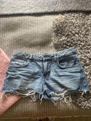 Blå jeansshorts med fransig kant - Säljer ett par blå jeansshorts med fransig och sliten kant. Klassisk femficksmodell med bälteshällor och dragkedja fram. Perfekta för sommaren och ger en avslappnad vibe.