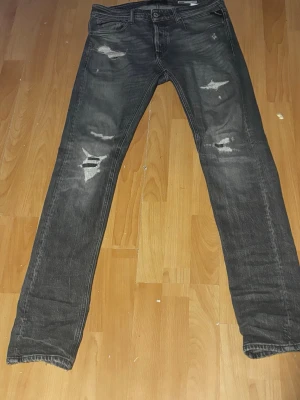 Svarta slitna jeans från Replay - Säljer ett par svarta jeans från Replay med snygga slitningar och hål på benen. Jeansen har fem fickor, raka ben och klassisk knappgylf. Perfekta för en cool och avslappnad stil. Materialet är denim i bomull.