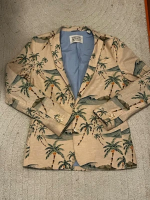 Beige kavaj med palmmotiv Scotch & Soda - Unik beige kavaj från Scotch & Soda med coolt palmmotiv och vågor i grönt och blått. Kavajen har ljusblått innerfoder, två knappar framtill och dekorativa knappar vid ärmsluten. Perfekt för dig som vill sticka ut med en tropisk vibe.