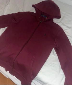 Vinröd hoodie från Polo Ralph Lauren - Säljer en vinröd hoodie från Polo Ralph Lauren med dragkedja framtill och klassisk huva. Hoodien har fickor på magen och en liten broderad logga på bröstet. Perfekt för en chill och stilren look.