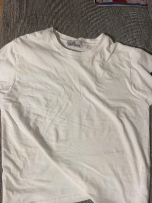 Vit t-shirt från Stone Island - Klassisk vit t-shirt från Stone Island med rund hals och korta ärmar. Tillverkad i mjukt bomullstyg och har den ikoniska Stone Island-loggan som avtagbar patch på ärmen. Perfekt för dig som gillar stilrena och trendiga plagg.