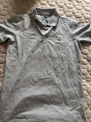 Grå pikétröja från Moncler - Snygg grå pikétröja från Moncler med klassisk krage och knappar framtill. Tröjan har Moncler-logga på bröstet och kragen har röd och blå randig detalj. Perfekt till en stilren och avslappnad look.