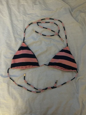 Randig trekantsbikini i rosa och svart - Säljer en snygg trekantsbikini med breda rosa och svarta ränder. Bikinin har knytband med guldfärgade detaljer och klassisk trekantsform. Perfekt för stranden eller poolen. Materialet känns som syntet, snabbtorkande och bekvämt.